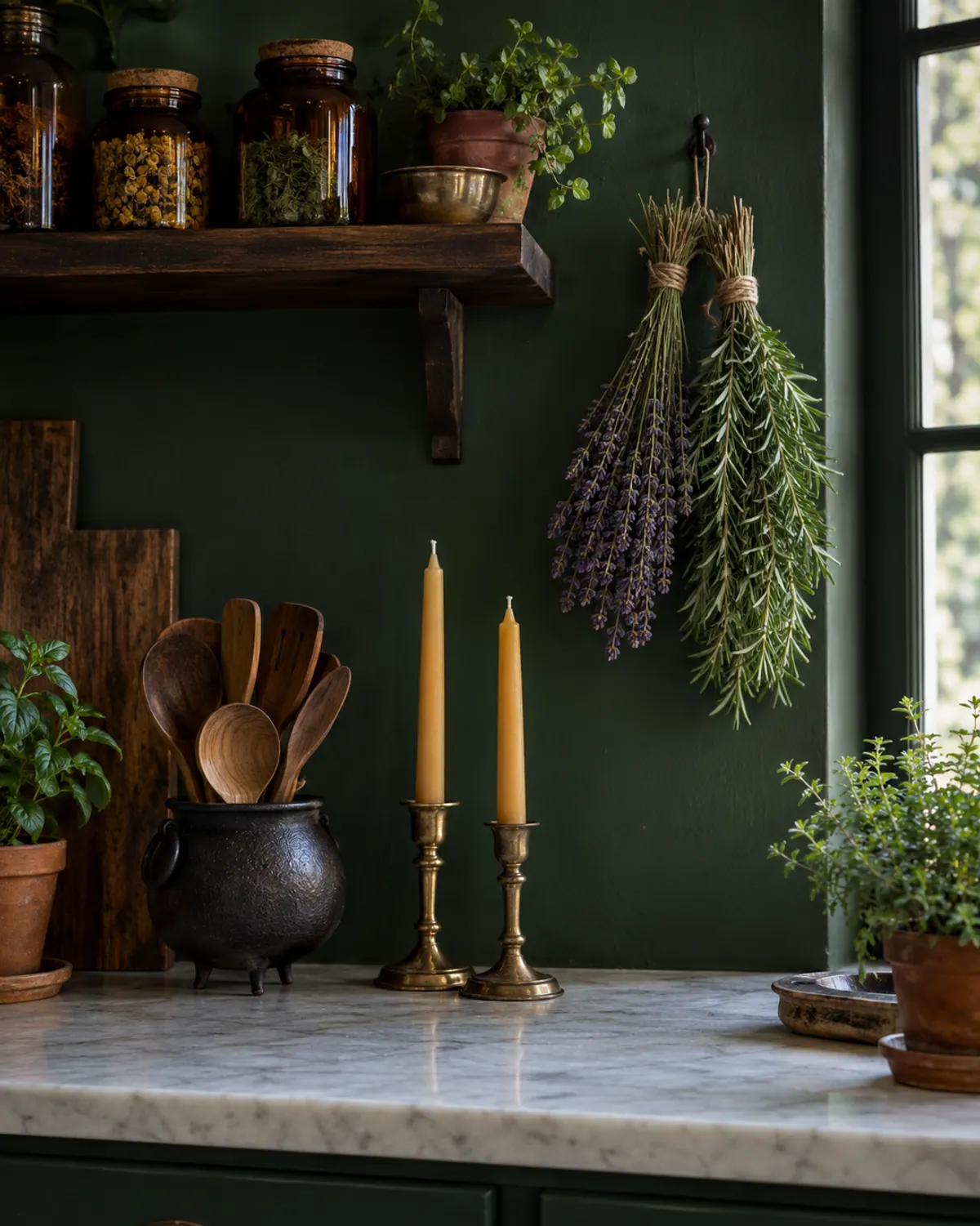 Witchy apothecary kitchen with a dark green wall, a wood shelf of amber jars, hanging lavender and rosemary bundles and brass candlesticks on a marble counter
